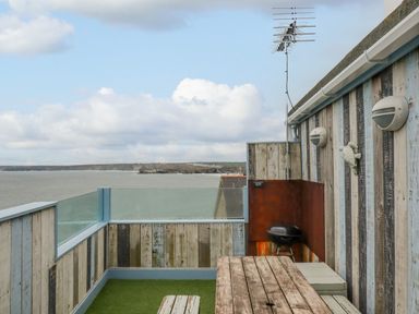 A rooftop patio with wooden walls glass railing a wooden table benches a barbecue grill and a view of the sea at Sailor's Rest in Newquay