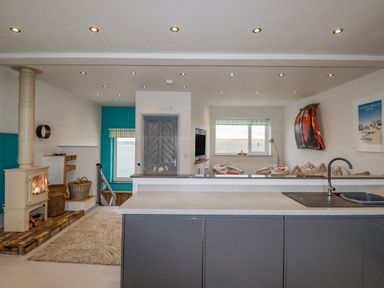 An open living space with a kitchen island a wood stove and a seating area with windows showing an ocean view at Sailor's Rest in Newquay