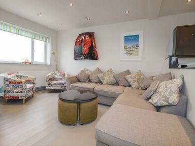A living room with a beige sectional sofa and two armchairs with text patterns a round table and wall art at Sailor's Rest in Newquay