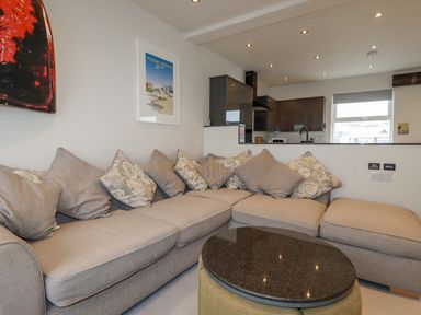 A living area with a sectional sofa and cushions next to a kitchen with cabinets and a window at Sailor's Rest in Newquay