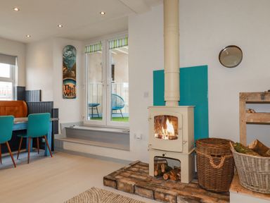 A room with a wood burning stove on a brick hearth two wicker baskets two turquoise chairs and a dining table at Sailor's Rest in Newquay