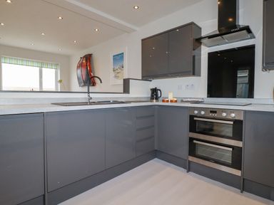 A modern kitchen with grey cabinets a built-in double oven a sink and a window with striped blinds at Sailor's Rest in Newquay