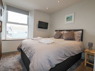A bedroom with a bed pillows folded towels a small wooden side table lamp a wall-mounted television and a window at Sailor's Rest in Newquay
