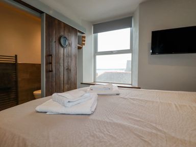A bedroom with a bed and folded towels near a window with a roller blind a wooden sliding door and a wall-mounted television at Sailor's Rest in Newquay
