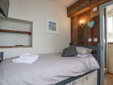 A bedroom with a single bed with gray bedding and a folded towel on it a wooden wall with a light and a heart decoration and a door on the side at Sailor's Rest in Newquay