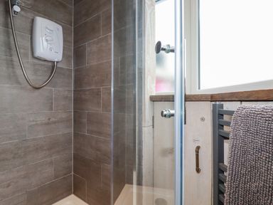 A shower area with brown tiled walls a white shower unit and a glass door next to a window and a towel rack with a gray towel at Sailor's Rest in Newquay