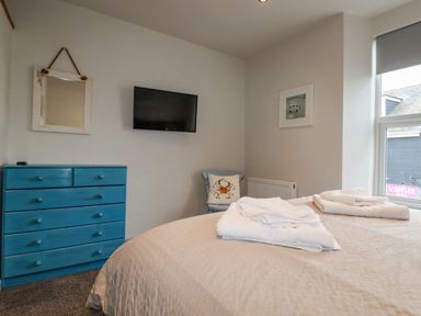 A bedroom with a bed, a blue chest of drawers, a wall-mounted TV, a chair with a cushion, and a window at Sailor's Rest in Newquay