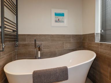 A bathroom with a freestanding bathtub a gray towel on the tub a wall-mounted radiator and a window with blinds at Sailor's Rest in Newquay