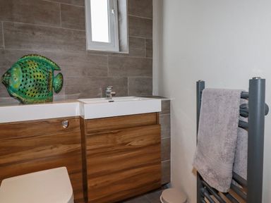 A bathroom with a wooden cabinet and sink a green fish decoration a gray towel on a towel rack and a small window at Sailor's Rest in Newquay