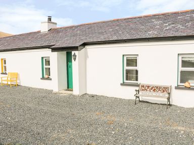 Exterior of a white cottage with green door and windows with two benches in a gravel yard at Pat White's Cottage in Rostrevor