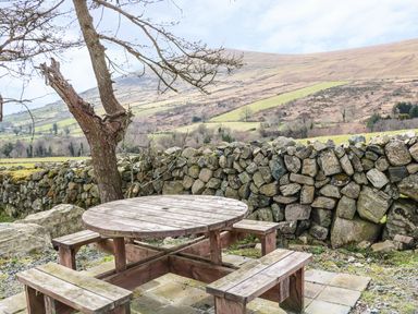 A wooden picnic table with benches on a tiled patio next to a stone wall and tree with hills in the background at Pat White's Cottage in Rostrevor