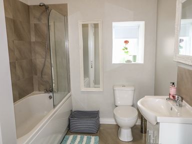 A bathroom with a bathtub shower combination toilet sink mirror window with a rose and woven basket on the floor at Pat White's Cottage Rostrevor