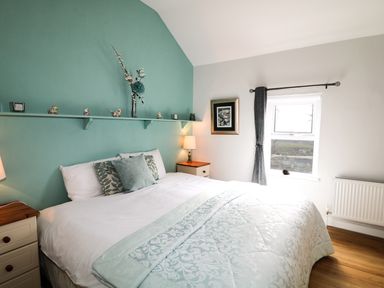 A bedroom with a double bed with pillows and a light blue patterned comforter and a window with curtain at Pat White's Cottage in Rostrevor