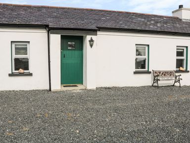 A white cottage exterior with a green door two windows a bench and gravel ground at Pat Whites Cottage in Rostrevor