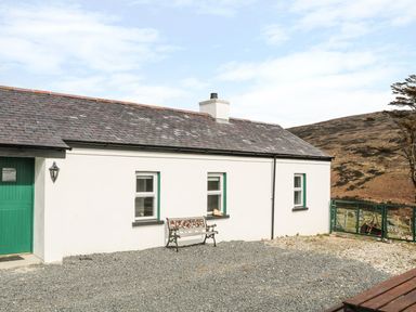 A white cottage with a green door and three windows with a bench outside on gravel at Pat White's Cottage in Rostrevor