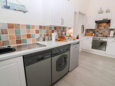 A kitchen with white cabinets, a stainless steel oven, washing machine, dishwasher, sink, toaster with bread, and tiled backsplash at Beachcomber in Marazion