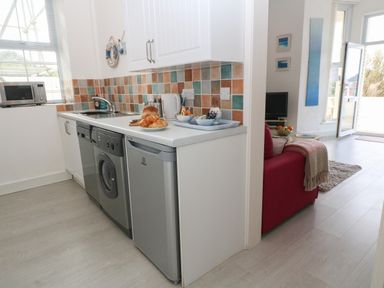 A kitchen with a microwave sink washing machine and mini fridge next to a living area with a red sofa and open door at Beachcomber in Marazion