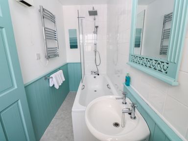 A bathroom with a bathtub shower combination a sink a mirror a towel rack and light blue paneling at Beachcomber in Marazion