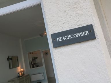 A sign reading Beachcomber on a textured exterior wall with a view into a room with a dresser a lamp and a kitchen area at Beachcomber Marazion