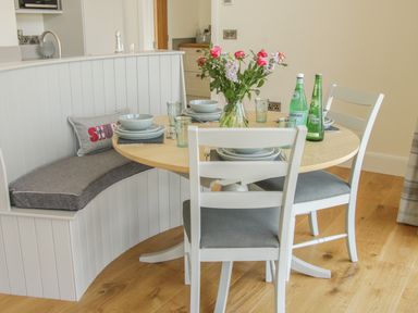 A dining area with a round wooden table set with bowls plates glasses and cutlery two white chairs with grey cushions and a curved bench with grey cushions and a pillow with the word snugs a vase with flowers and two bottles of sparkling water at Shepherd's Rest in Montgomery