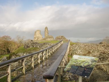 A wooden bridge leading to ruins of a stone castle with an information sign at a historical site in Montgomery