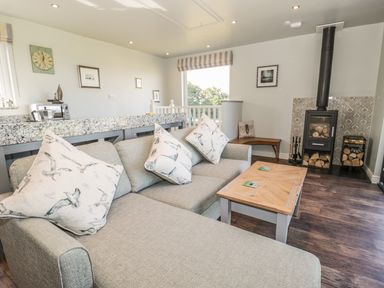 A living room with a grey sofa with bird patterned cushions a wooden coffee table a wood burning stove with logs and a granite kitchen counter at Emblestones Embleton