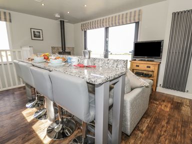 A kitchen with granite countertop and four gray stools next to a gray sofa and a wooden TV stand with a TV at Emblestones in Embleton