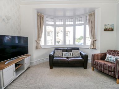 A living room with a leather sofa and a striped armchair facing a large bay window and a television on a wooden cabinet at 20 Ulwell Road in Swanage
