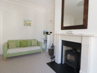 A room with a green sofa with cushions a fireplace with a wooden framed mirror above and a small white shelf with books at 20 Ulwell Road in Swanage