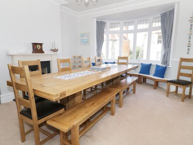 A dining room with a large wooden table surrounded by wooden chairs and benches with a bay window seating area with cushions at 20 Ulwell Road in Swanage
