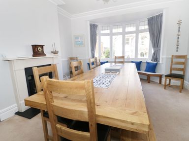A dining room with a wooden table and chairs a fireplace and a bay window with cushions at 20 Ulwell Road in Swanage