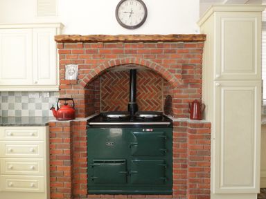 A kitchen area with a green Aga stove set in a red brick alcove with a clock on the wall and cream cabinets on either side at 20 Ulwell Road in Swanage