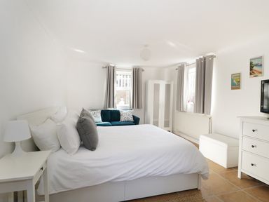 A bedroom with a white bed and pillows a bedside table with a lamp a teal sofa two windows with gray curtains a white wardrobe a white dresser with a TV and framed pictures on the wall at 20 Ulwell Road in Swanage
