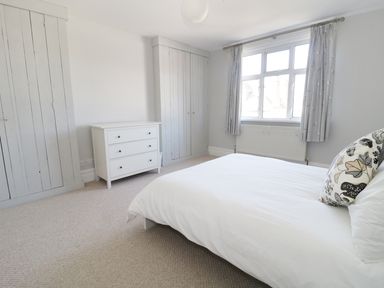 A bedroom with a bed white dresser built in wardrobes and a window with curtains at 20 Ulwell Road in Swanage
