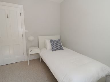 A small bedroom with a single bed with white bedding and a black and white cushion a white bedside table with a lamp and a closed white door at 20 Ulwell Road in Swanage