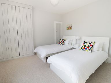 A bedroom with two single beds with white linens and colorful pillows a white wardrobe and a small white bedside table at 20 Ulwell Road Swanage