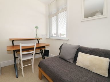 A small room with a wooden desk and chair next to a window and a gray sofa with two cushions at 20 Ulwell Road in Swanage