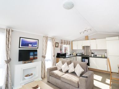 A living room with a sofa TV and kitchen area with cabinets at 32 Cruachan Lodge in Auchterarder