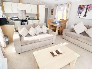 A living area with beige sofas a wooden coffee table a dining table with chairs and a kitchen with white cabinets at 32 Cruachan Lodge in Auchterarder