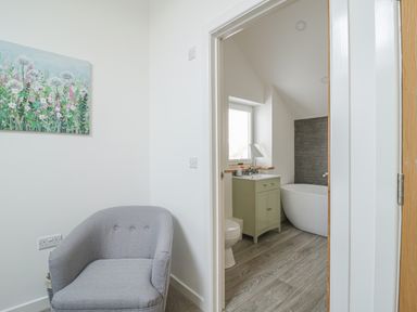 A gray armchair next to a wall with floral painting and a view into a bathroom with a bathtub sink cabinet and toilet at Coney Garth Barn in Beckermet