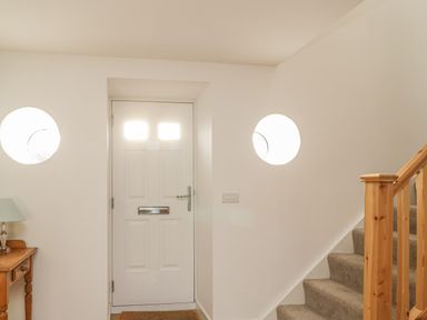 An entryway with a white front door two round windows a wooden side table with a lamp and stairs with a wooden banister at Coney Garth Barn in Beckermet