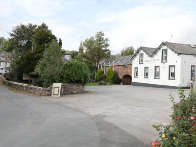 A parking lot next to a building with white walls and black trim with sign white mare bar restaurant and a road with stone wall and trees
