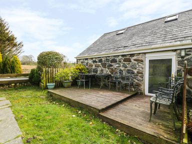 An outdoor wooden deck with metal chairs and tables next to a stone house at Bwthyn Ael Y Bryn in Criccieth