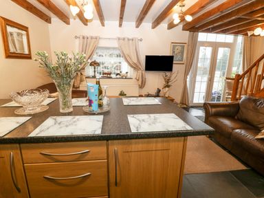 A kitchen island with placemats and flowers with a living area featuring a leather sofa and a wall-mounted TV at Bwthyn Ael Y Bryn in Criccieth