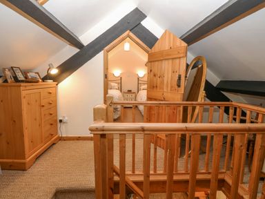 A wooden staircase and railing leading to a small landing with a chest of drawers lamp and photos and an open wooden door showing a bedroom with two beds at Bwthyn Ael Y Bryn in Criccieth