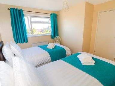 A bedroom with two single beds with white linens teal runners and folded towels by a window with teal curtains at Howards Hill West in Cromer
