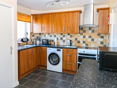 A kitchen with wooden cabinets a washing machine a stove a microwave and a window with orange blinds at Howards Hill West in Cromer