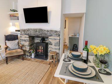 A living room with a stone fireplace stove a mounted TV an armchair and a dining table set with dishes wine and flowers at Llys Madoc in Porthmadog