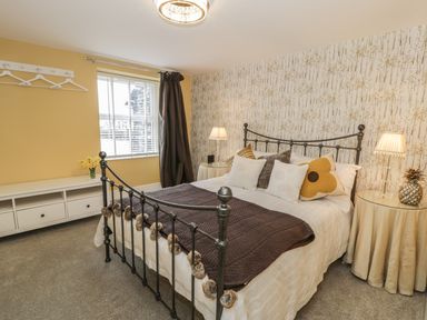 A bedroom with a metal bed frame a bed with pillows and a brown throw two round bedside tables with lamps and decorations a window with blinds and dark curtains a white storage unit with drawers and yellow walls at Llys Madoc in Porthmadog