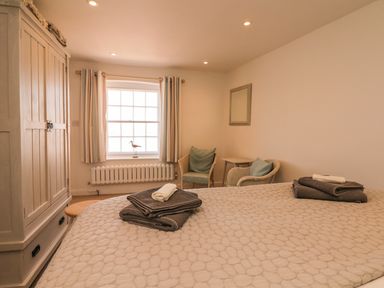 A bedroom with a bed covered with a quilt towels on the bed a wooden wardrobe two chairs a small table and a window with curtains at Searles in Lympstone